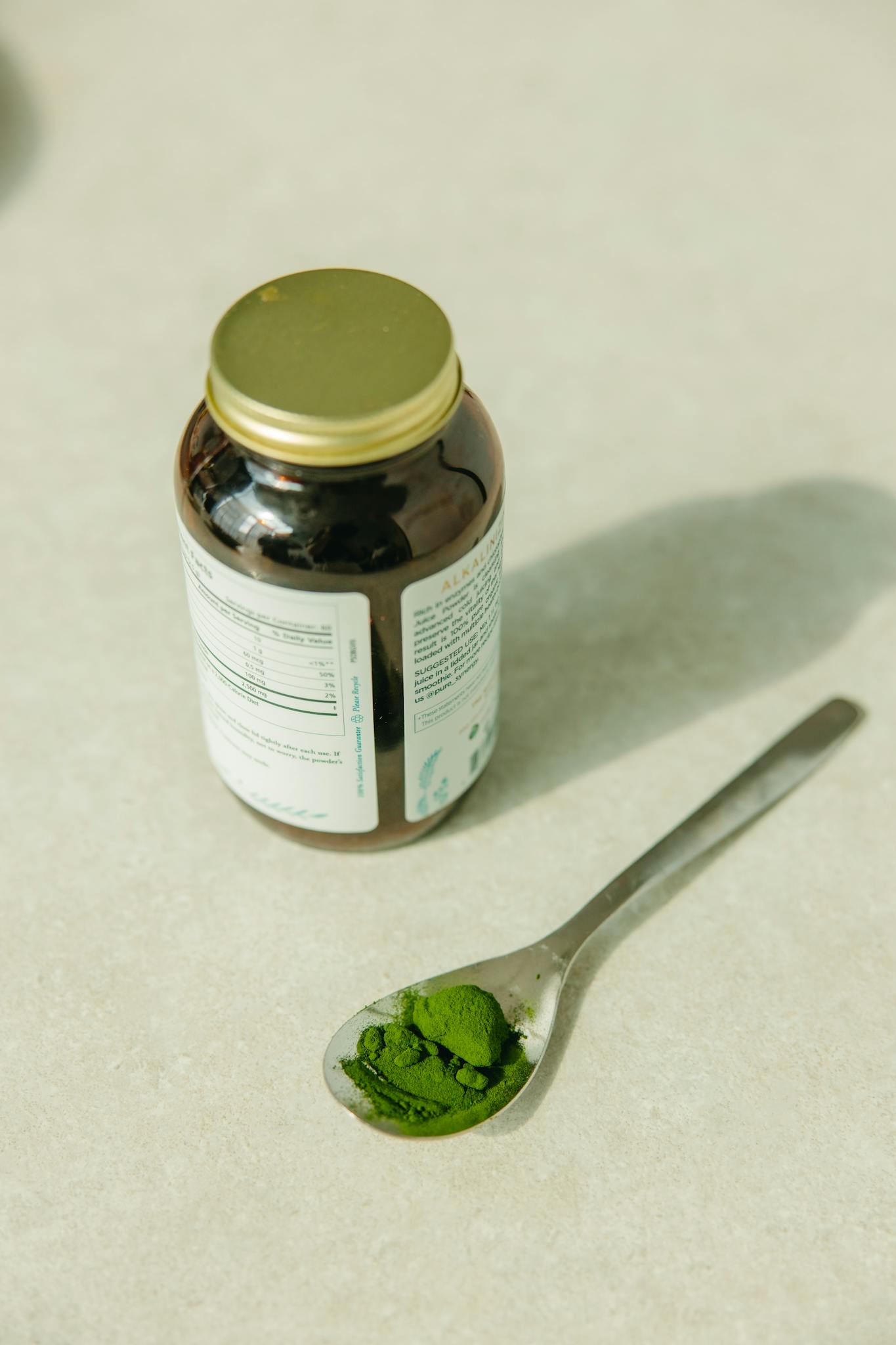 Top view of green supplement powder in a spoon next to a glass jar on a neutral background.